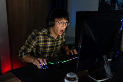 Asiatischer Teenager mit aufgeregtem Gesichtsausdruck beim Spielen eines PC-Spiels in einem abgedunkelten Raum